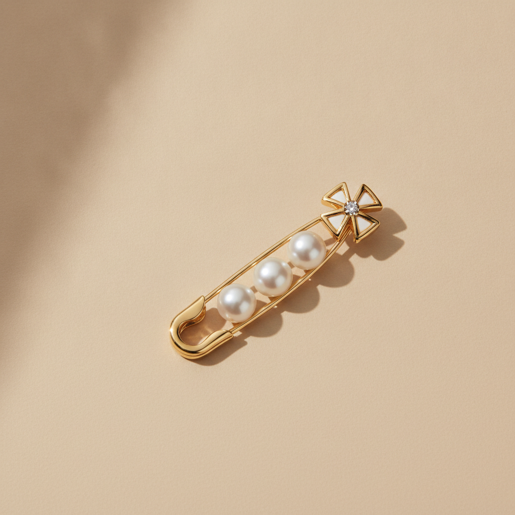 Illustration: a gold safety pin-shaped brooch featuring three white pearls and a decorative top with white enamel and a small diamond, resting on a beige surface